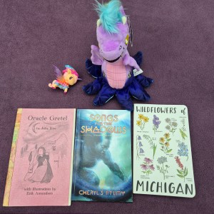 Photo of a small unicorn, a purple dragon plushie with tentacles, a chapbook titled "Oracle Gretel", a copy of novella "Songs for the Shadows", and a notebook with "Wildflowers of Michigan".